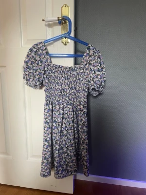 Blommig kortklänning från Missguided - Supersöt blommig kortklänning från Missguided Petite med puffärmar och smockad överdel. Klänningen har lila, blå och vita toner med små blommor och en figurnära passform upptill. Perfekt för sommarens alla tillfällen!