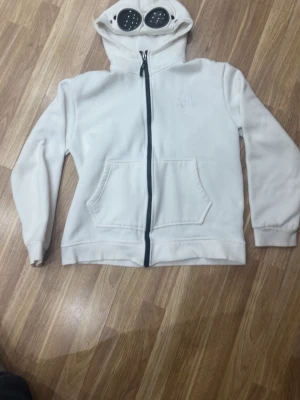 Vit hoodie med goggles från C.P. Company - Vit hoodie  C.P. Company. Ikonisk huva med inbyggda goggles och broderad logga på bröstet. Stora framfickor och svarta detaljer vid dragkedjan. Perfekt för dig som vill sticka ut med streetwear-stil.