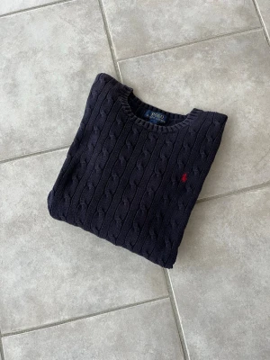 Mörkblå kabelstickad tröja Polo Ralph Lauren - Snygg mörkblå kabelstickad tröja från Polo Ralph Lauren med rund halsringning och röd broderad logga på bröstet. Tröjan har långa ärmar och ribbade muddar vid ärmslut och nederkant. Perfekt för lager på lager och klassisk stil.
