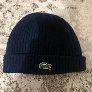 Mörkblå ribbstickad mössa Lacoste - Snygg mörkblå ribbstickad mössa från Lacoste med klassisk krokodillogga framtill. Tillverkad i 100% ull för extra värme och komfort. Enkel design med uppvikt kant, perfekt för kalla dagar och streetstyle.