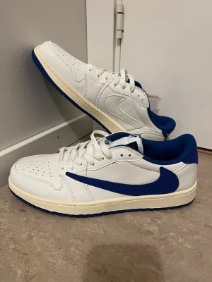 Nike Jordan 1 Low vit/blå sneakers - Nike Jordan 1 Low sneakers i vitt och blått med klassisk Swoosh i blått på sidan. Skorna har en rund tå, snörning och en blå yttersula med bra grepp. Ovandelen är i slätt skinn och innersulan är mjukt vadderad. Medföljer extra skosnören i rosa och blått.