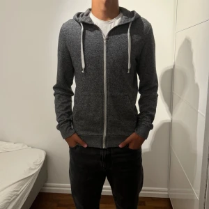 Mörkgrå hoodie med dragkedja - Mörkgrå hoodie med vit dragkedja och snören. Klassisk modell med huva, kängurufickor och ribbade muddar. Perfekt för chill dagar eller när du vill ha en enkel och stilren look. Materialet är mjukt och känns skönt mot huden.