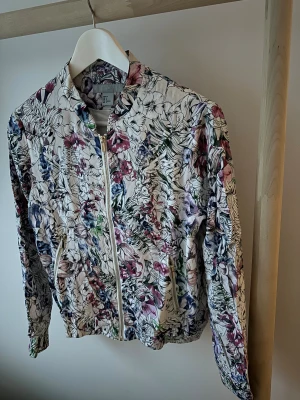 Blommig bomberjacka från H&M, strl 36 - Snygg bomberjacka från H&M i storlek 36 med blommigt mönster i lila, blått, grönt och rosa på ljus botten. Jackan har dragkedja framtill, två fickor med dragkedja och resår i midja och ärmslut. 