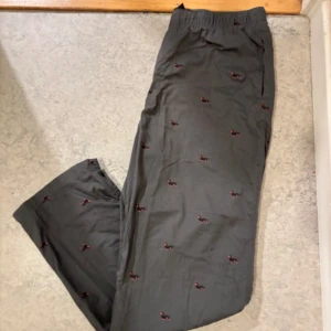 Svarta pyjamasbyxor med hundmönster - Svarta byxor med elastisk midja och fickor, dekorerade med små broderade taxar i rött och svart över hela tyget. Byxorna har rak passform och är perfekta för chill dagar hemma eller när du vill ha något unikt.