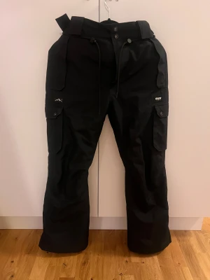 Svarta täckbyxor från Dope  - Säljer ett par svarta täckbyxor från Dope Snow i storlek S. Cargopants inspirerande, med lite bootcut form. Använt under en vintersäsong, annars perfekt skick. Pris kan förhandlas. 