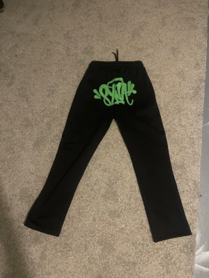 Syna world  - Svarta mjukisbyxor med elastisk midja och snörning. På baksidan finns en stor grön graffiti-inspirerad print och på ena benet står det 'Forgive Me' i grönt. Byxorna har rak passform och är gjorda i mjukt material, perfekta för chill dagar.