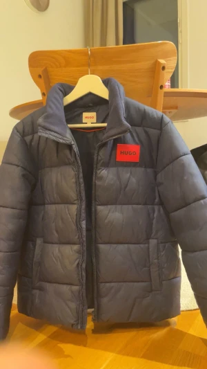 Mörkblå pufferjacka från HUGO - Snygg mörkblå pufferjacka från HUGO med röd logopatch på bröstet. Jackan har quiltad design, dragkedja framtill och mjukt foder i kragen. Perfekt för kalla dagar och har en clean, modern look som funkar till streetwear. Köpt föraåret för 2000