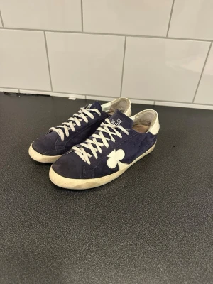 The Gallerist skor - Snygga mörkblå sneakers från The Gallerist med vit snörning och vit klöverdetalj på sidan. Skorna är i mocka med vit häl och platt sula. Dem är väldigt använda och inte i så bra skick därför priset. Priset är ej hugget i sten så de är bara höra av sig vid frågor eller funderingar.