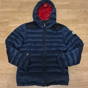 Mörkblå dunjacka från Moncler - Snygg mörkblå dunjacka från Moncler med röd insida på huvan. Jackan har horisontella quiltade paneler, dragkedja framtill och två sidofickor med dragkedja. Perfekt för kalla dagar och har en modern look med huva och Moncler-logga på ärmen.