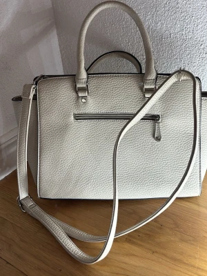 Ljusgrå handväska med axelrem och dragkedja - Snygg  handväska i skinnimitation med både handtag och justerbar axelrem. Väskan har en stilren, boxig form och en yta med struktur. Utsidan har en dragkedja och insidan är fodrad med blommigt mönster samt flera fack för smart organisering.