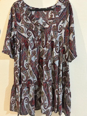 Mönstrad kortklänning med paisley - Kortklänning med paisleyinspirerat mönster i rött, blått, svart och grönt på vit botten. Klänningen har v-ringning, korta ärmar och en lös, flowy passform med volangdetaljer. Perfekt för dig som gillar färg och mönster.