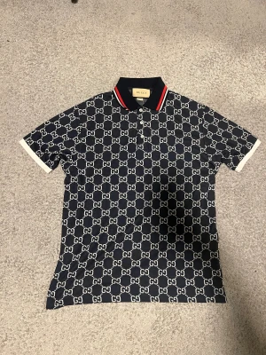 Mörkblå GG-mönstrad piké  - Exklusiv mörkblå pikétröja med ikoniskt vitt GG-mönster över hela plagget. Kragen är svart med röda och vita ränder och knäppning framtill med fyra vita knappar. Pikén har korta ärmar med vita muddar och är tillverkad i ett bekvämt bomullsmaterial.