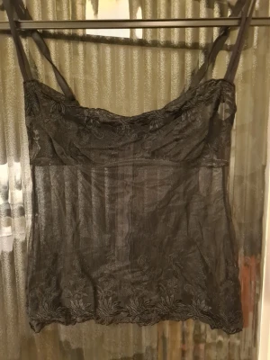 Svart spetsig linnetopp med blommönster - Säljer en svart, transparent linnetopp med tunna justerbara axelband och detaljerad spets med blommönster längs överdelen och nederkanten. Klaspar i ryggen, ingen stretch