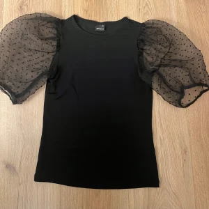 Svart topp med puffärmar från Gina Tricot - Säljer en svart topp från Gina Tricot i storlek XS med transparenta puffärmar i mesh och prickigt mönster. Perfekt för dig som vill ha en cool och trendig look med extra volym på ärmarna.