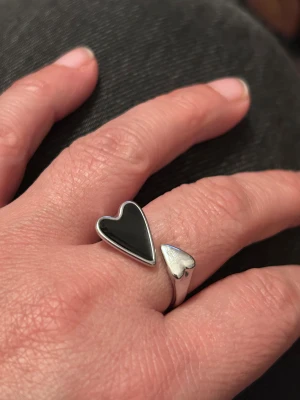 Silverring med svarta och silvriga hjärtan - Stål silverring med två hjärtan, ett större svart och ett mindre i blankt silver. Ringen har en öppen design som gör den justerbar och ger en cool, modern vibe. Perfekt för dig som gillar statement-smycken med personlig touch.