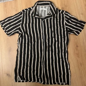 Randig skjorta från S.C.W. i svart och beige - Snygg kortärmad skjorta från S.C.W. i svart med beige och vita ränder. Skjortan har klassisk krage och knäppning framtill. Perfekt för dig som gillar en chill och stilren look. Materialet känns lätt och skönt mot huden.