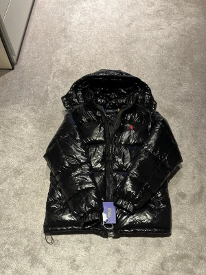 Svart pufferjacka från Polo Ralph Lauren - Svart glansig pufferjacka från Polo Ralph Lauren med röd broderad logga på bröstet. Jackan har huva, dragkedja framtill och elastiska muddar vid ärmslut. Perfekt för kalla dagar och har en riktigt snygg streetstil.