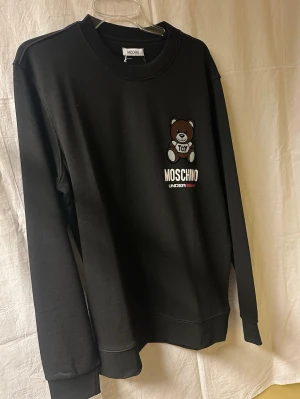 Svart Moschino sweatshirt med björn - Svart sweatshirt från Moschino med rund hals och lång ärm. På bröstet finns ett broderat motiv av en brun nalle med texten 'TOY' samt Moschino Underbear-logga i vitt och rött. Tröjan är gjord i mjuk bomull och har en clean, normal passform.