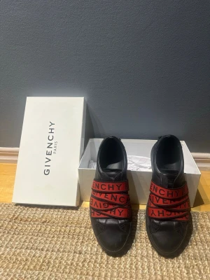Givenchy svarta sneakers med röda band - Säljer ett par svarta Givenchy sneakers i skinn med breda röda elastiska band över foten där Givenchy-loggan syns tydligt. Klassisk rund tå och platt sula. Kommer med originalkartong och dustbag. Riktigt snygga och trendiga skor för dig som gillar exklusiva detaljer.