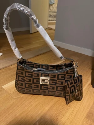 Brun och svart axelväska från Guess - Snygg axelväska från Guess med grafiskt G-mönster i brunt och svart. Väskan har guldfärgade detaljer, dragkedja upptill och en matchande liten plånbok som hänger i en kedja. Stilren form och logoplatta framtill.
