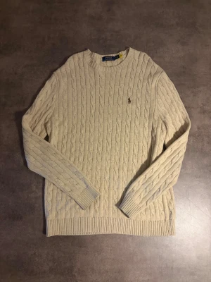Beige kabelstickad tröja Polo Ralph Lauren - Säljer en beige kabelstickad tröja från Polo Ralph Lauren med klassisk rund hals och broderad logga på bröstet. Tröjan har ribbade muddar och är långärmad, perfekt för dig som gillar stilren och tidlös design.