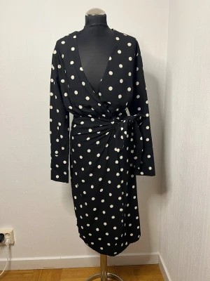 Svart prickig omlottklänning H&M XS - Svart omlottklänning från H&M i storlek XS med vita prickar. Klänningen har lång ärm, knyt i midjan och v-ringad hals. Den är knälång och har en mjuk, lätt känsla som gör den skön att bära. Perfekt för dig som gillar retrovibbar och tydliga kontraster. Aldrig blivit använd men utan prislapp. 