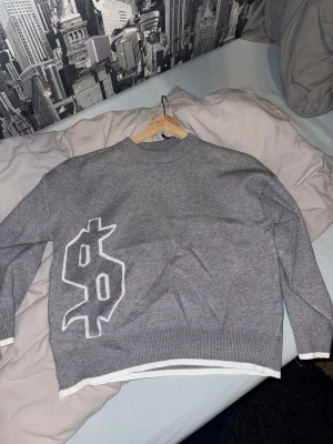 Grå stickad tröja med tryck - Säljer en grå stickad tröja med vit dollartryck på framsidan och texten 'buy happiness' i vit gotisk stil på ryggen. Tröjan har ribbade muddar och nederkant med vit detalj, samt långärmad passform och rund hals. Perfekt för dig som gillar streetwear och statement-plagg.