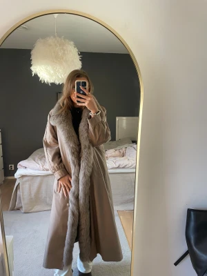 Beige lång kappa med pälsdetaljer - Säljer en beige lång kappa med bred krage och framkant i fluffig gråbrun päls. Insidan är fodrad med mörkbrun päls, vilket gör den extra varm. Jackan har en loose passform och långa ärmar, perfekt för dig som vill ha en statement piece i garderoben. Pälsen som sitter på kragen är troligtvis oäkta. 