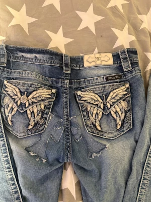 Miss Me jeans med broderade fickor, strl 27 - Snygga blå jeans från Miss Me med midwaist och slim bootcut. Baksidan har detaljerade broderier och glittriga stenar på fickorna, med coola vingmotiv. Jeansen har slitningar och kontrastsömmar.🌟❣️ kan sänka pris!