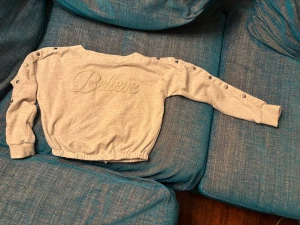 Glittrig beige tröja från Believe XXS - Snygg beige tröja från Believe med glittriga detaljer och texten 'Believe' broderad framtill. Tröjan har långa ärmar med tryckknappar längs axlar och ärmar samt ribbade muddar och elastisk nederkant. Perfekt för dig som gillar unika detaljer. Mycket bra skick förutom ett litet hål på insidan (fån) som syns på en av bilderna och kan lätt åtgärdas 