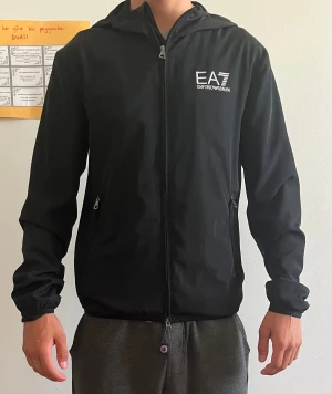 Svart EA7 vindjacka med huva - Svart vindjacka från EA7 Emporio Armani med huva och vita logodetaljer på bröstet. Jackan har dragkedja framtill och på fickorna, samt meshfoder på insidan. Perfekt för dig som gillar stilren och sportig look.
