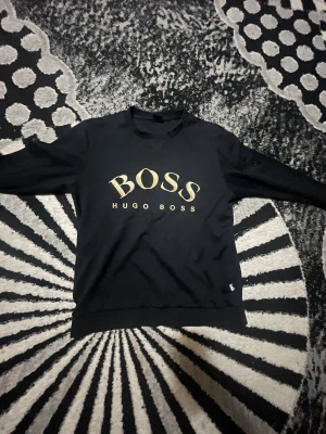 Svart sweatshirt från Hugo Boss - Säljer en svart sweatshirt från Hugo Boss med stort guldfärgat BOSS-tryck på bröstet. Tröjan har rund halsringning och långa ärmar. Perfekt för dig som gillar stilrena och klassiska plagg med en lyxig touch.