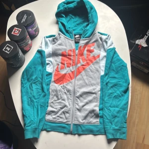Nike hoodie i turkos och grå  - Nike hoodie i turkos och ljusgrått med stor orange logga framtill. Tröjan har dragkedja, huva och långa ärmar. Skön standard fit och perfekt för chill eller träning. Materialet är mjukt och känns som bomullsmix. Skick 10/10 använts 2 gånger i 2023.  Pris kan diskuteras
