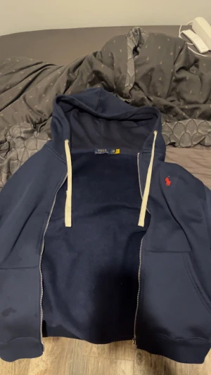 Marinblå hoodie från Polo Ralph Lauren - Snygg marinblå hoodie från Polo Ralph Lauren med dragkedja och vit snörning i huvan. Tröjan har ett rött Polo-logo broderat på bröstet och är tillverkad i mjuk bomull. Perfekt för chill dagar och har långa ärmar samt praktiska fickor framtill.