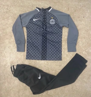 Grå långärmad PSG x Nike träningströja - Snygg långärmad träningströja från Nike i samarbete med PSG. Framsidan har ett geometriskt mönster i mörkgrått och svart, med PSG-logga och Qatar Airways-tryck. Ärmarna är enfärgat grå och materialet känns lätt och sportigt, perfekt för fotboll eller träning.