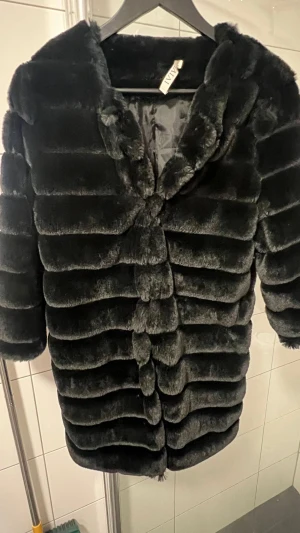 Svart pälsjacka från IVIVI - Säljer en svart pälsjacka från IVIVI med horisontella ränder och mjuk, fluffig känsla. Jackan har en rak form, trekvartsärmar och är fodrad på insidan. Perfekt för dig som vill ha en lyxig och varm look under kalla dagar.