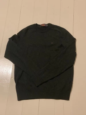 Morris stickad tröja merinoull - Snygg mörkgrå stickad tröja från Morris i 100% merinoull. Rund hals och diskret broderad logga på bröstet. Har krymp lite i storleken. Den har långa ärmar och ribbade muddar. Perfekt för dig som gillar stilrena och bekväma plagg.