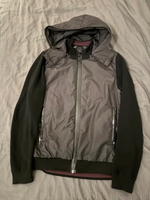 Hugo Boss x Porsche Cardigan - Svart cardigan från Boss x Porsche med huva och hel dragkedja framtill. Jackan har släta ärmar i stickad material och framstycke i syntet, samt två sidofickor med dragkedja. Perfekt för dig som gillar stilren och sportig look. Nypris ungefär 2500