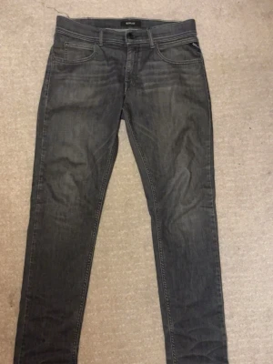 Replay grå jeans slim fit - Snygga grå jeans från Replay med klassisk femficksdesign och diskreta slitningar. Modellen har smal passform och raka ben, med tydliga sömmar och Replay-logga vid fickan. Perfekt för dig som gillar stilrena och moderna jeans.