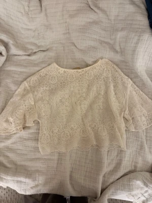 Vit spetsblus med vida ärmar - Superfin vit blus i spets med vackert blommigt mönster och vida, korta ärmar. Croppad modell med rund halsringning och romantiska detaljer. Perfekt för dig som vill ha en söt och luftig topp med extra detaljer.