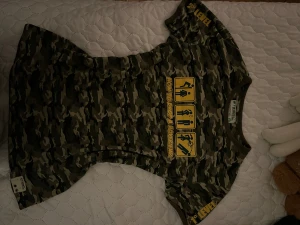 Kamouflage t-shirt med tryck från Stfood - Cool t-shirt i grönt kamouflagemönster med gul text och grafik på bröstet: 'How to drop a boyfriend' och roliga symboler. På axeln står det 'LEVEL' och på ryggen 'GAME OVER'. Rund hals och korta ärmar, perfekt för dig som gillar edgy streetstyle.