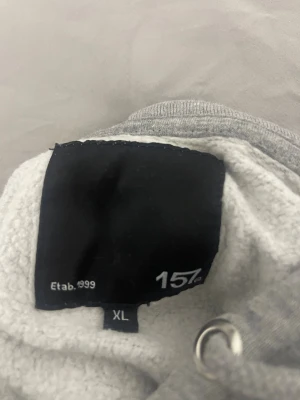 Grå hoodie från 157 med snörning - En enkel och clean grå hoodie från 157 i storlek XL. Tröjan har huva med snörning och en klassisk magficka. Mjuk insida och ribbade muddar vid ärmslut och nederkant. Perfekt för chill dagar eller när du vill ha något bekvämt.