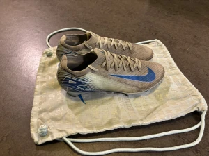 Nike Mercurial Air Zoom Elite fotbollsskor ljusblåa  - Säljer ett par Nike Mercurial Air Zoom Elite fotbollsskor i ljusblått med blå detaljer och Swoosh. Skorna har snörning, låg profil och en rund tå. Ovandelen är tillverkad i syntetmaterial med mönstrade linjer på sidan. Perfekt för dig som vill ha grym touch och snabb känsla på planen då skorna knappt väger någonting och är väldigt lätta.
