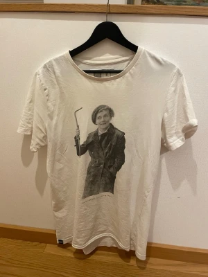 Vit t-shirt med tryck från Dedicated - Snygg vit t-shirt från Dedicated i 100% ekologisk och Fairtrade-certifierad bomull. Framsidan har ett coolt svartvitt tryck av en person i trenchcoat med en krok i handen(Astrid Lindgren). Klassisk rund hals och korta ärmar. Perfekt för dig som gillar grafiska motiv.