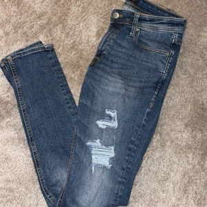 Jack & Jones jeans  - Säljer ett par blå jeans med slim passform och snygga slitningar på framsidan. Jeansen har klassisk femficksdesign, knapp och dragkedja i midjan samt kontrastsömmar. Perfekt för dig som gillar en cool och avslappnad stil.