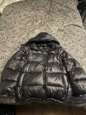 Svart pufferjacka från Polo Ralph Lauren - Svart pufferjacka från Polo Ralph Lauren med huva och två stora fickor med dragkedja. Jackan har en röd Polo-logga på bröstet och quiltad design. Perfekt för kalla dagar med sin tjocka och fluffiga look. Storleken är S