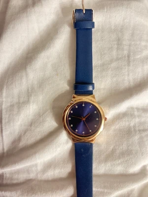 Blå klocka med guldfärgad boett - Stilren klocka med blått läderband och matchande blå urtavla. Boetten är guldfärgad och urtavlan har små stenar som timmarkeringar. Enkel och elegant design som passar till många outfits.