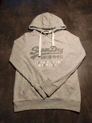Superdry hoodie - Tvärfet superdry hoodie med unikt glitrande loga, denna är en klassisk hoodie som passar perfekt till alla årstider. Hör av dig vid frågor!