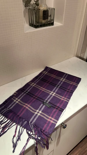 Lila rutig halsduk från Burberry - Säljer en lila rutig halsduk från Burberry i 100% lammull. Halsduken har klassiskt rutmönster i lila, rosa, svart och beige samt fransar i ändarna. Perfekt accessoar för att lyfta din outfit.