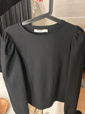 Svart ribbad långärmad topp NA-KD - Säljer en svart ribbad topp från NA-KD i storlek S. Toppen har puffade axlar och långa ärmar, vilket ger en snygg siluett. Materialet är mjukt och stretchigt, perfekt för dig som gillar stilrena och enkla plagg med lite edge.